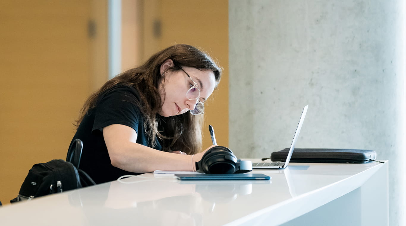 A woman sitting at a table takes notes while a laptop computer is open in front of her