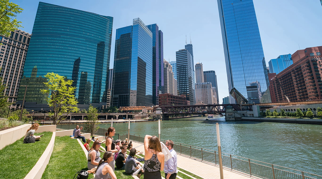 View of high-rise buildings along the Chicago River in downtown Chicago, on a sunny day. Friends gather nearby at a green park on the river's edge.