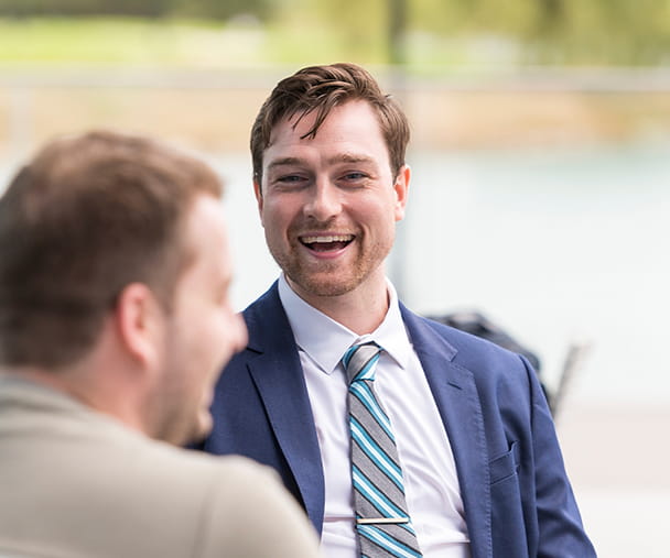 A Kellogg student in a navy suit and striped tie sits outside on a bench and talks with his classmate