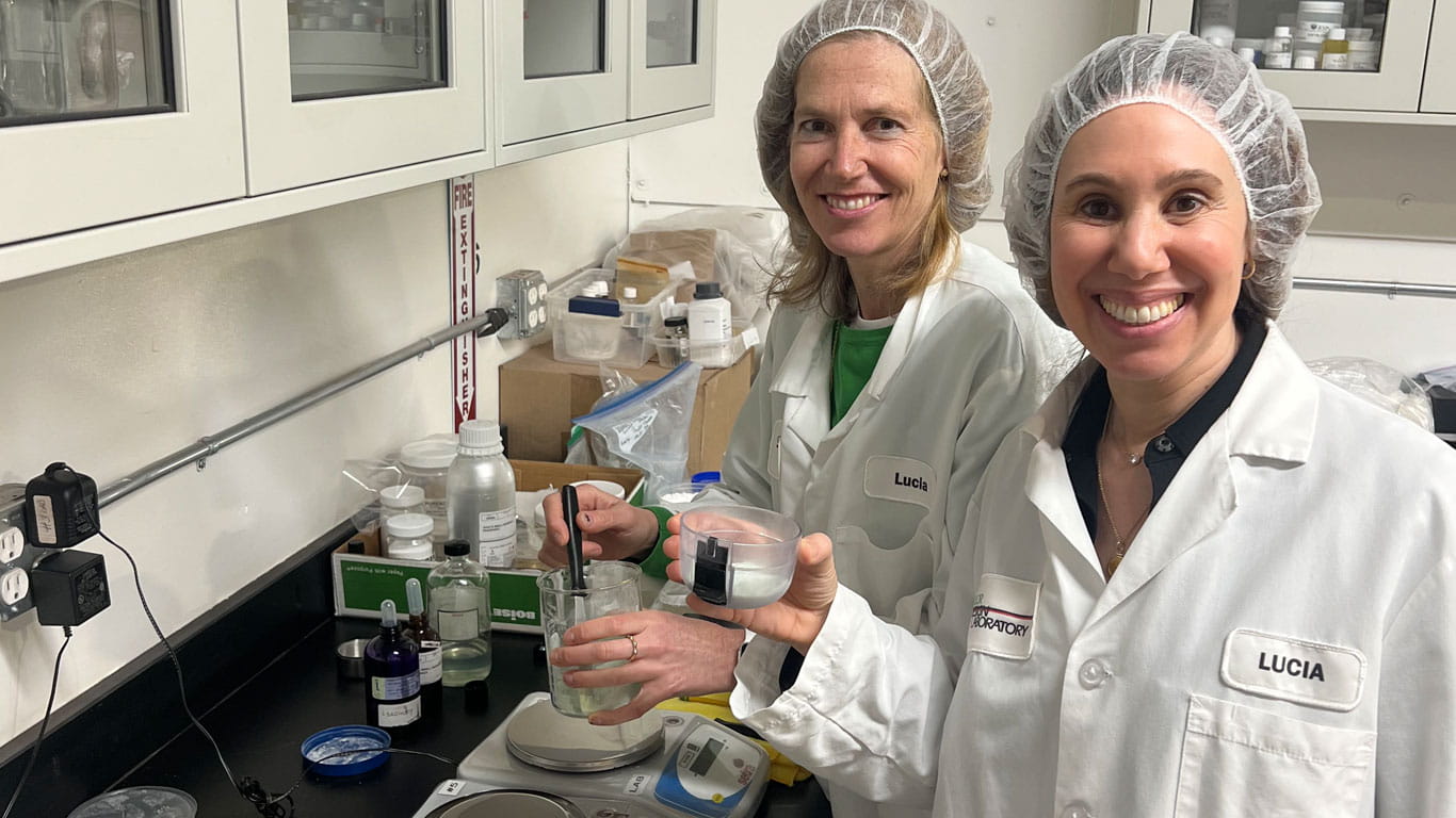 Small Wonder co-founders Stephanie Farsht and Katja Lerner stand in front of a chemistry lab workbench, wearing white lab coats and hairnets as Katja formulates their powdered shampoo product.