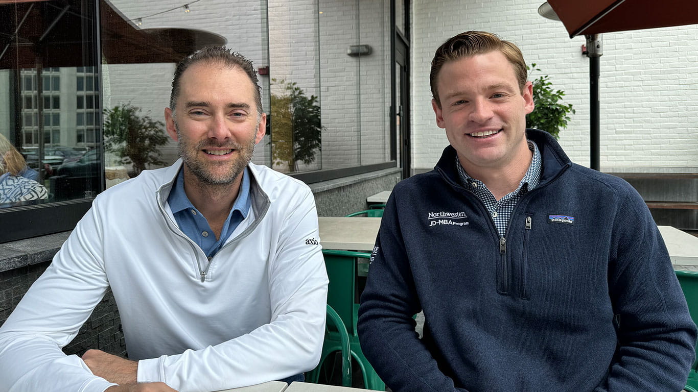 Axio's Scott Kannry and Sam Skinner, both JD-MBA grads, sit side by side at a table. Sam wears a dark pullover with the Kellogg JD-MBA Program logo.