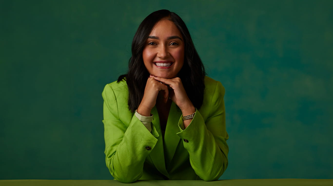 Yaritza Vallejo poses for a photo, resting her chin on her hands and smiling. She wears a light green blouse and sits in front of a dark green background.