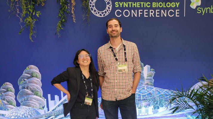 Opera Bioscience cofounders Julie Ming Liang and Gerry Sapienza stand in front of a poster of a futuristic illustration, with signage reading "Synthetic Biology Conference"