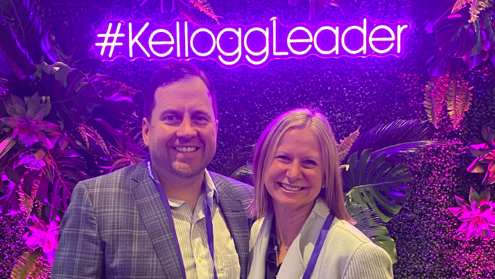 Kellogg faculty Katie Arnold and Evan Norton smile together in front of a neon purple sign reading "Kellogg Leader"