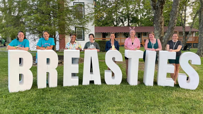 Alumna Lindsey Baker poses with fellow breast cancer survivors at Camp Breastie, behind a sign with large white letters spelling BREASTIES.