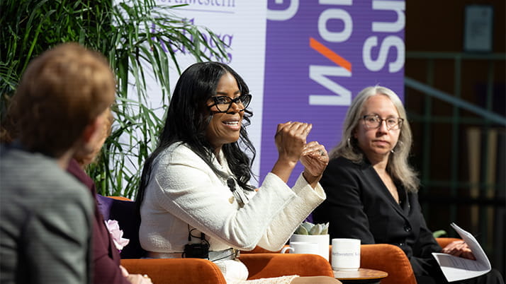 Three professional women participate in a panel discussion at the Kellogg Global Women's Summit. The speaker in the center, dressed in a white suit and gesturing expressively, addresses the audience with enthusiasm. To her left and right, the other panelists listen attentively. A purple 'Women's Summit' banner is visible in the background, and the setting is adorned with greenery and modern decor.