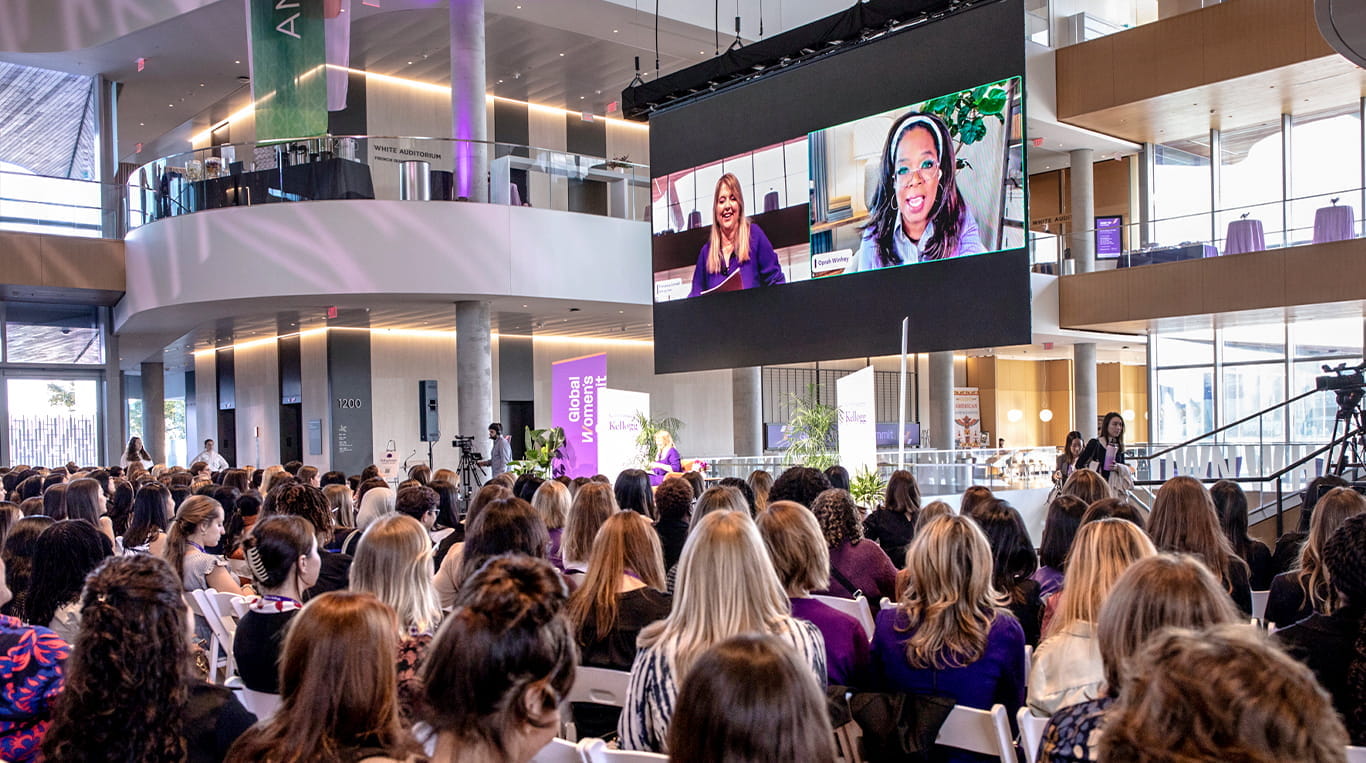 A large crowd gathers in Gies Plaza at the Global Hub at the Kellogg Global Women's Summit, attentively watching a virtual session displayed on a big screen. Oprah Winfrey and Dean Francesca Cornelli engage in a conversation projected for the audience. The space features high ceilings, glass walls, and purple-themed decor, creating a professional and inspiring atmosphere.