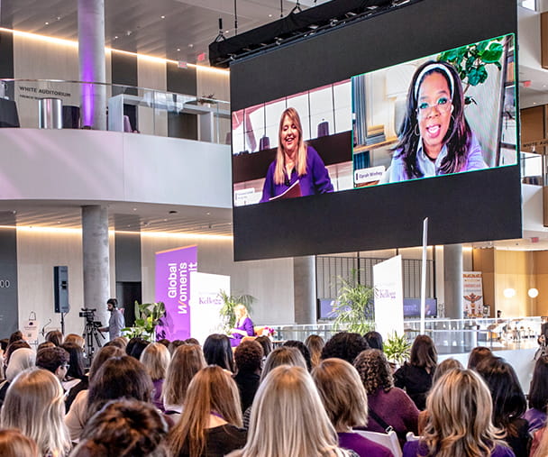 A large crowd gathers in Gies Plaza at the Global Hub at the Kellogg Global Women's Summit, attentively watching a virtual session displayed on a big screen. Oprah Winfrey and Dean Francesca Cornelli engage in a conversation projected for the audience. The space features high ceilings, glass walls, and purple-themed decor, creating a professional and inspiring atmosphere.