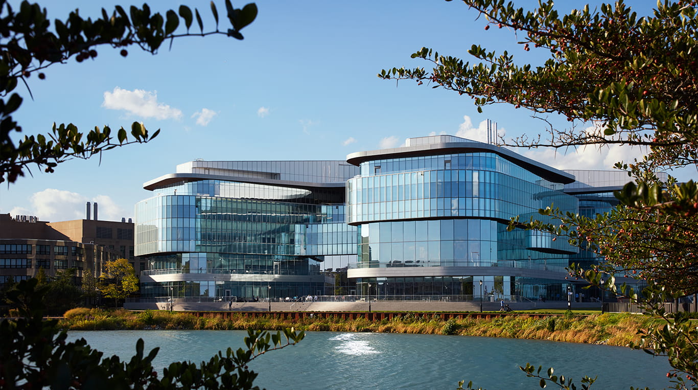 The Kellogg School of Management’s Global Hub building stands prominently along the water, its modern glass facade reflecting the clear blue sky. The striking architecture is framed by leafy tree branches, highlighting the serene and picturesque campus setting.