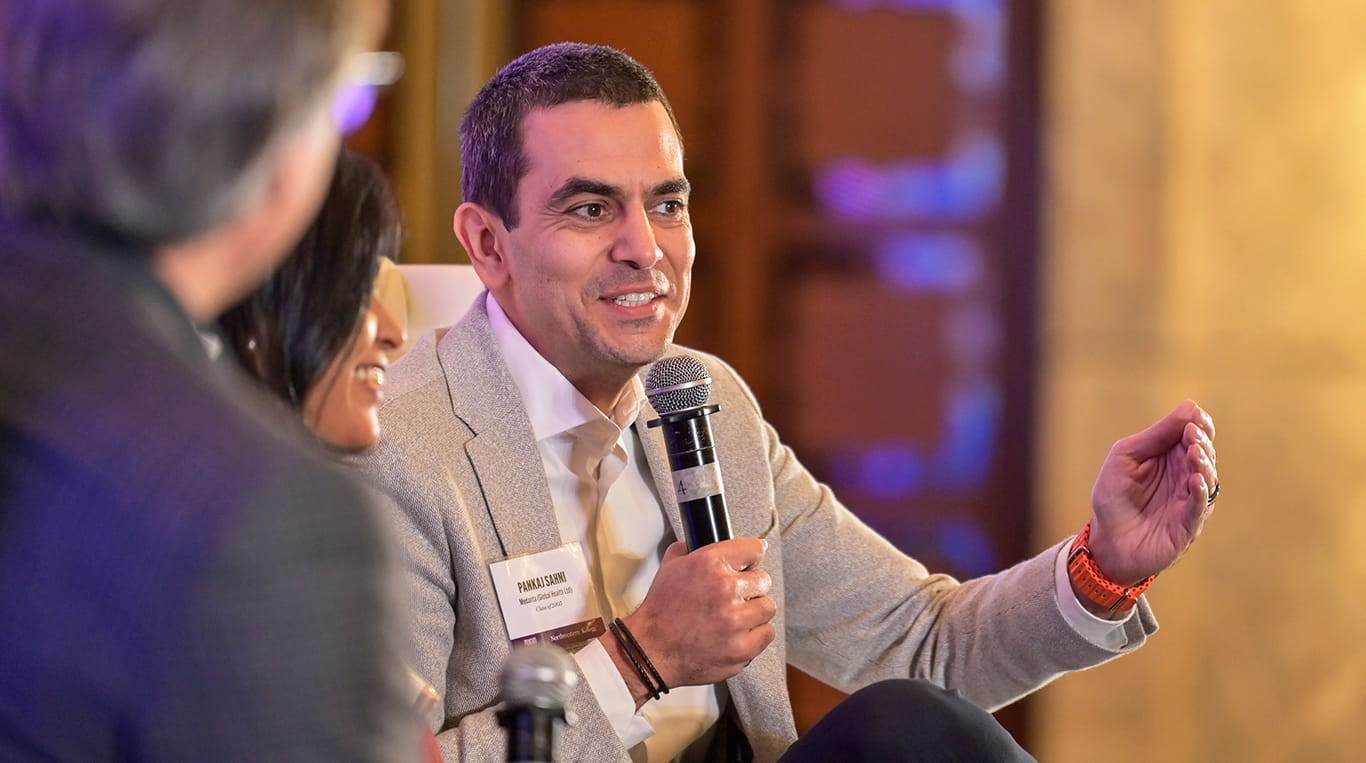 Medanta CEO Pankaj Sahni speaks at the Pragati conference in New Delhi. He wears a beige blazer and white shirt speaks into a microphone while gesturing with his hand during a panel discussion. wo other panelists, a woman and a man, are partially visible in the foreground, engaged in the conversation. The background features warm lighting and blurred decorative elements.