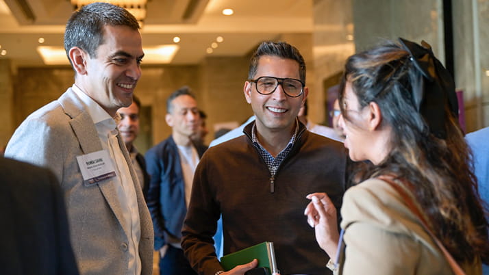 A group of people engaged in conversation at Pragati. On the left, a man in a light beige suit with a name badge is smiling while listening. In the center, a man wearing glasses and a brown sweater with a collared shirt underneath is smiling broadly. On the right, a woman with long dark hair partially pulled back is speaking animatedly with her hand raised in conversation.