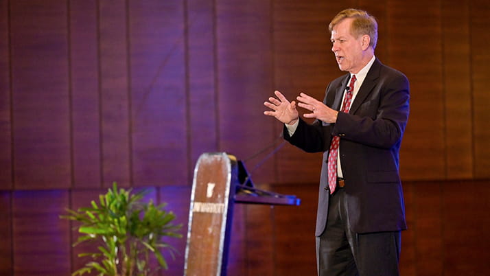 Professor Harry Kraemer presents at the Pragati conference. He wears a dark suit with a red patterned tie stands on stage gesturing with both hands while speaking. The stage has wooden paneled walls with purple lighting effects visible in the background.