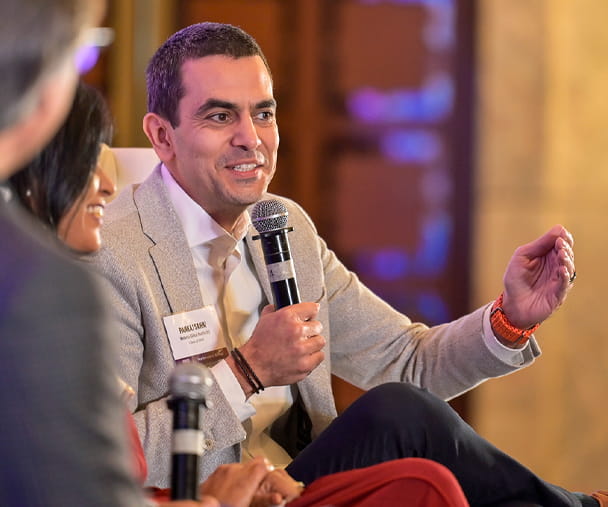 Medanta CEO Pankaj Sahni speaks at the Pragati conference in New Delhi. He wears a beige blazer and white shirt speaks into a microphone while gesturing with his hand during a panel discussion. wo other panelists, a woman and a man, are partially visible in the foreground, engaged in the conversation. The background features warm lighting and blurred decorative elements.