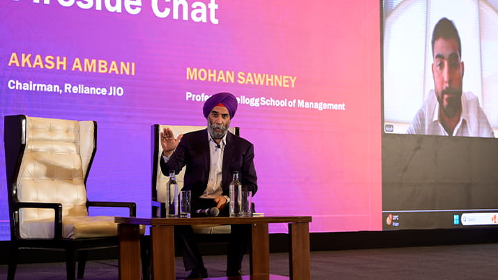 A fireside chat at the Pragati conference. On stage, Professor Mohan Sawhney wears a turban and dark suit and sits at a wooden table with water bottles, gesturing while speaking. Next to him is an empty cream-colored chair. A large screen displays a video feed of Reliance JIO chairman Akash Ambani joining remotely.