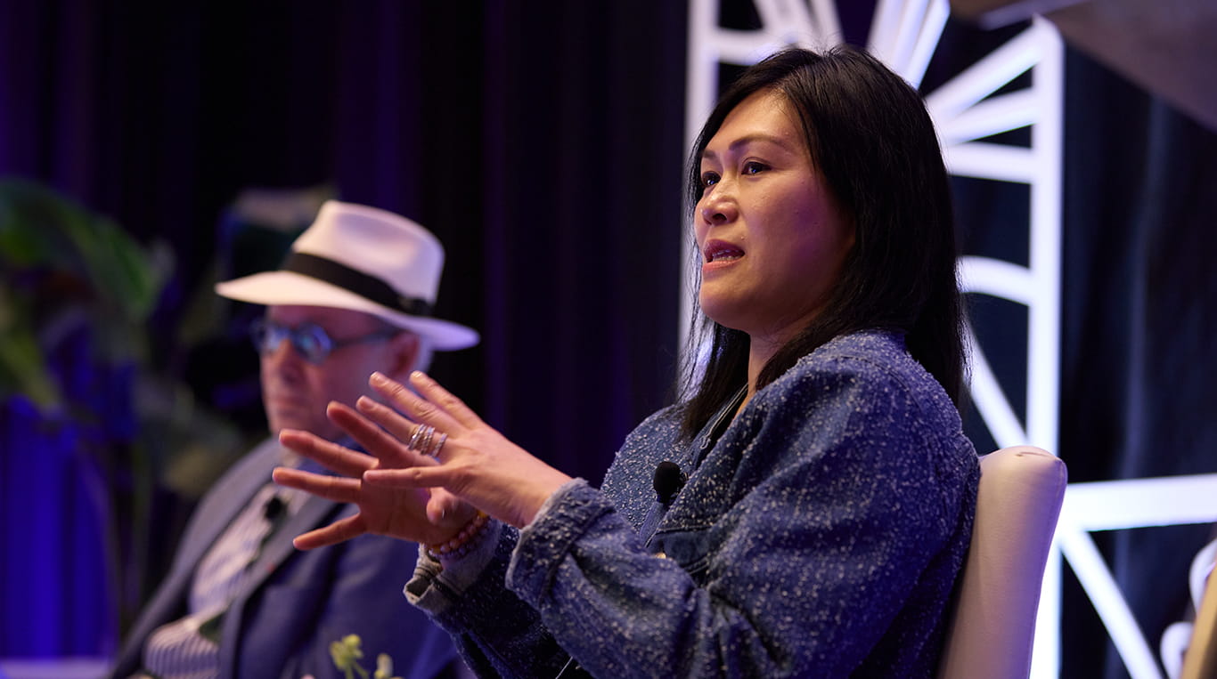 A woman in a blue jacket speaks passionately on stage, using expressive hand gestures to emphasize her points. She wears a microphone and sits against a visually striking backdrop with geometric designs and dramatic lighting. In the background, a man in a white hat and blue blazer listens attentively, contributing to the engaged and thoughtful atmosphere of the panel discussion.