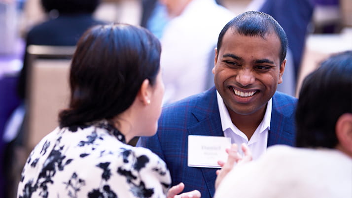 A man in a blue checkered suit jacket with a name badge is smiling broadly while engaged in conversation with a woman seen from behind. She has dark hair and is wearing a black and white patterned blouse or jacket. They are attending a conference at the Kellogg Miami Campus. The image captures a moment of genuine connection and positive interaction between the two individuals.
