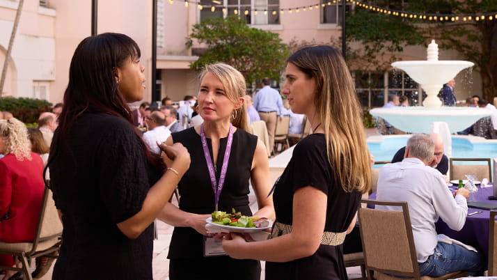 Three women standing and talking at an outdoor reception and networking event at the Kellogg Miami Campus. In the background is a decorative fountain, string lights overhead, and other attendees seated at tables. 