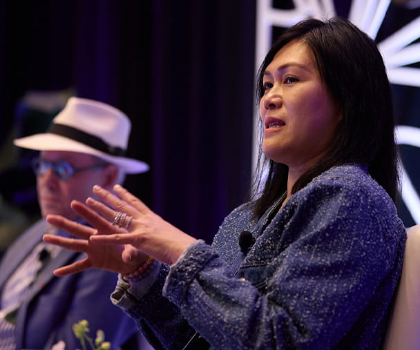 A woman in a blue jacket speaks passionately on stage, using expressive hand gestures to emphasize her points. She wears a microphone and sits against a visually striking backdrop with geometric designs and dramatic lighting. In the background, a man in a white hat and blue blazer listens attentively, contributing to the engaged and thoughtful atmosphere of the panel discussion.